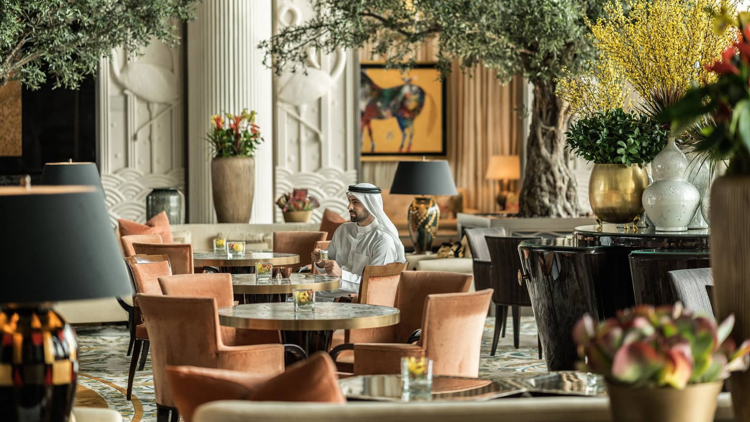 Man in traditional Bahraini dress sitting in lounge, surrounded by round tables with rust chairs and lush greenery