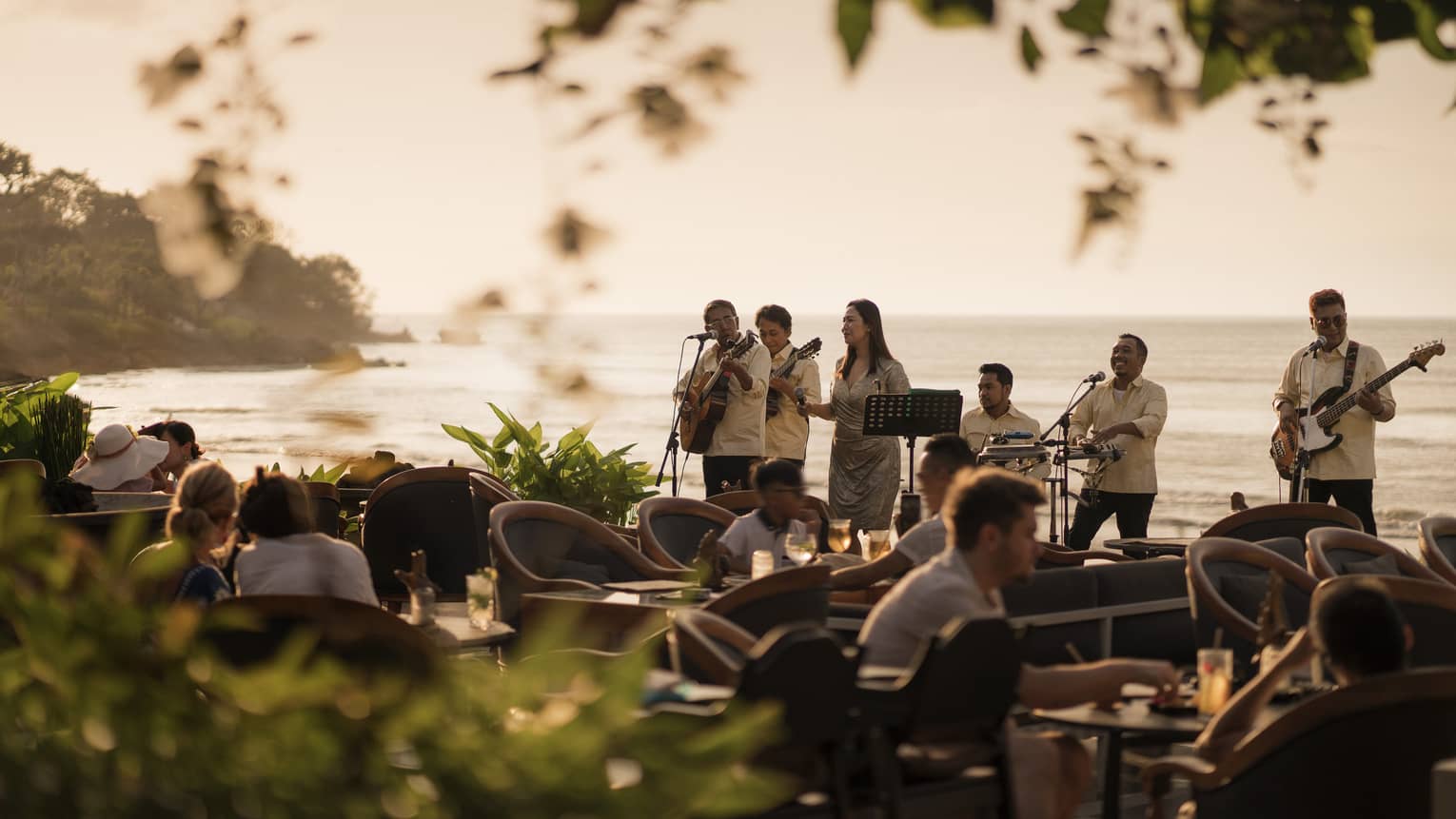 View through foliage of a musical band standing with their backs to the ocean and playing for dining guests at dusk.
