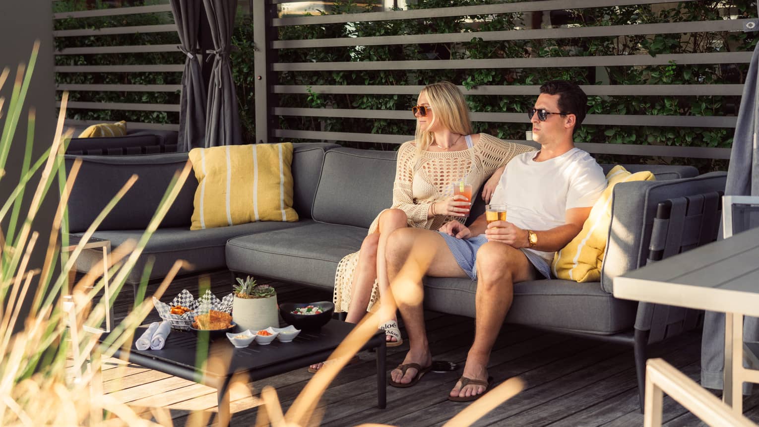 Two people sit on a patio couch with drinks, surrounded by cushions and snacks on a table. They wear sunglasses and casual summer attire.