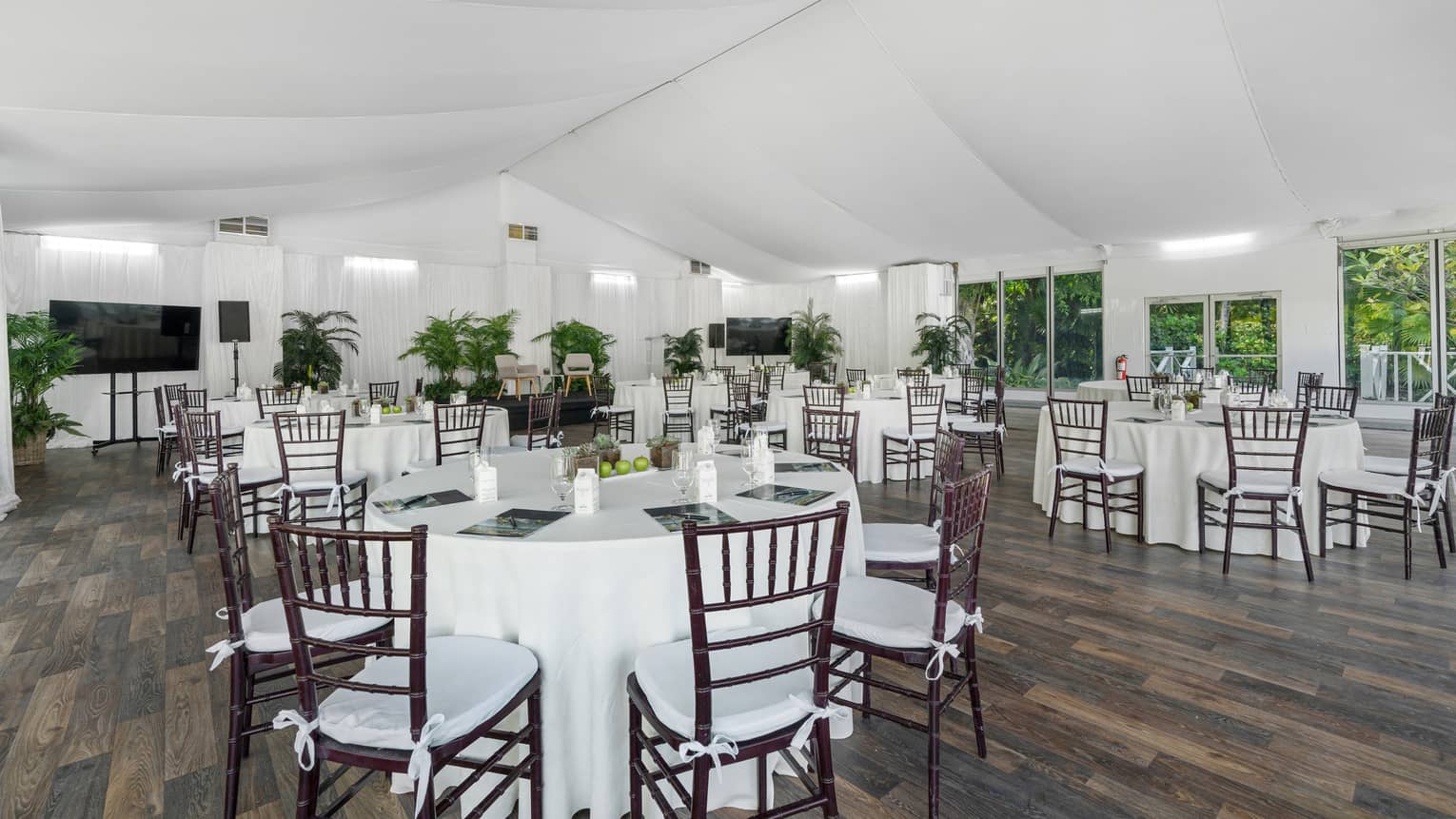 White event tent set up with dark wood flooring, six round tables furnished with brown cane chairs and various green plants lining one side wall with windows along another wall