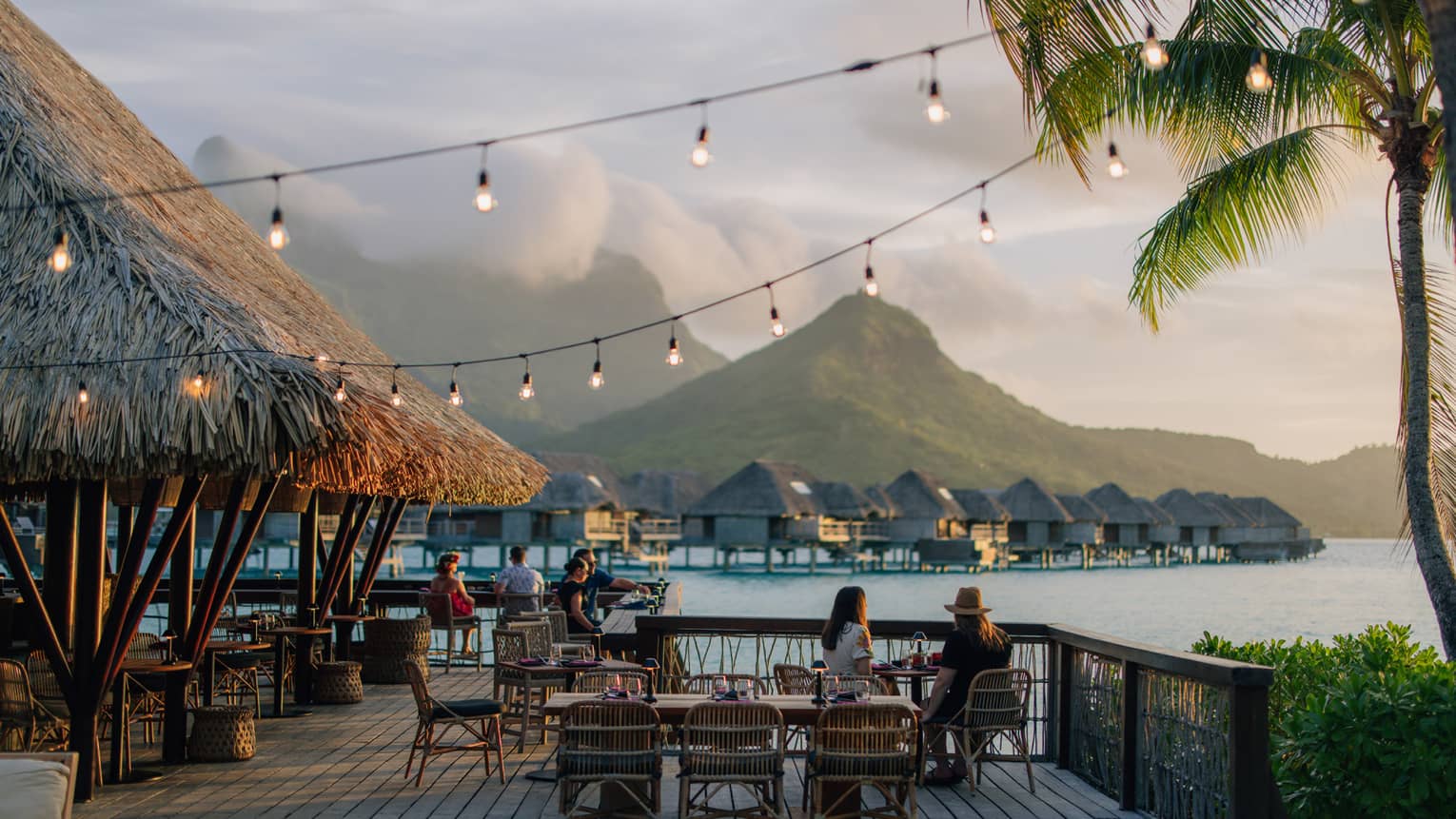 Groups of people dine outdoors on wooden deck, overwater pavilions and mountains in background