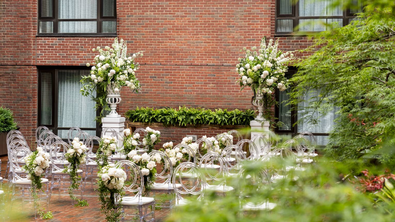 An outdoor wedding ceremony near a large red brick wall, with many rows of chairs and flowers.