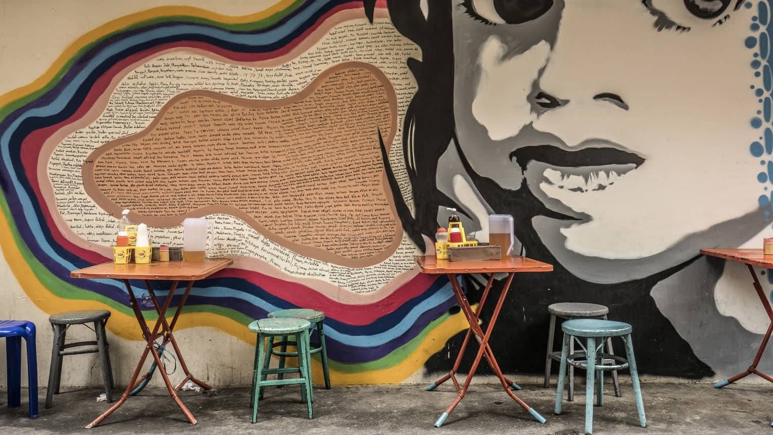 Street art mural featuring a large black-and-white face on a wall, with small, colourful dining tables and stools in front it