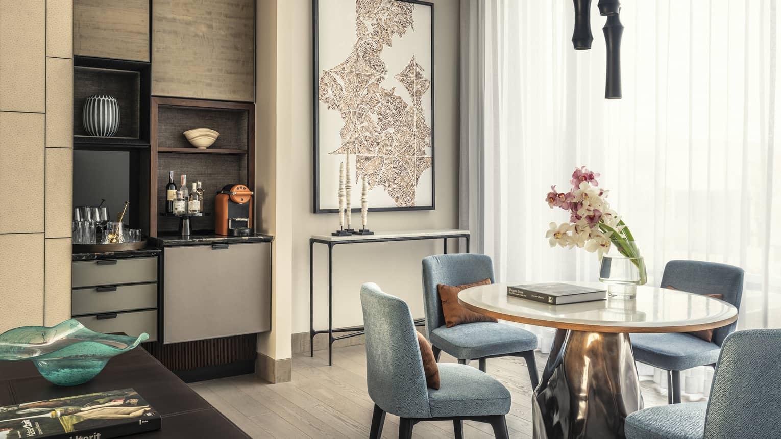Modern dining room with light blue chairs, a round white table, a vase of orchids and a small bar area with a coffee machine and liquor bottles. 