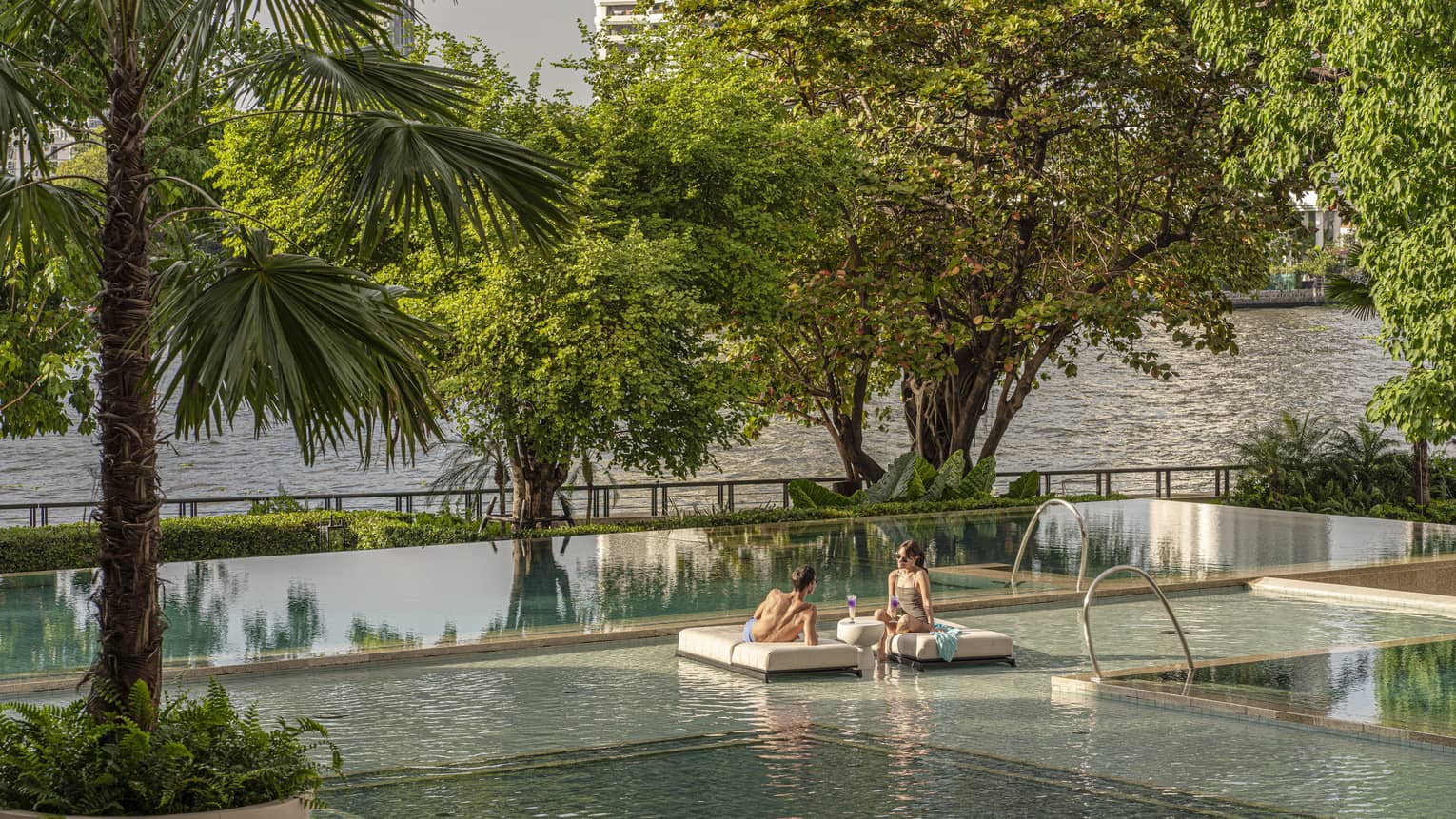 A man and woman lounging on small benches floating on a pool outside.