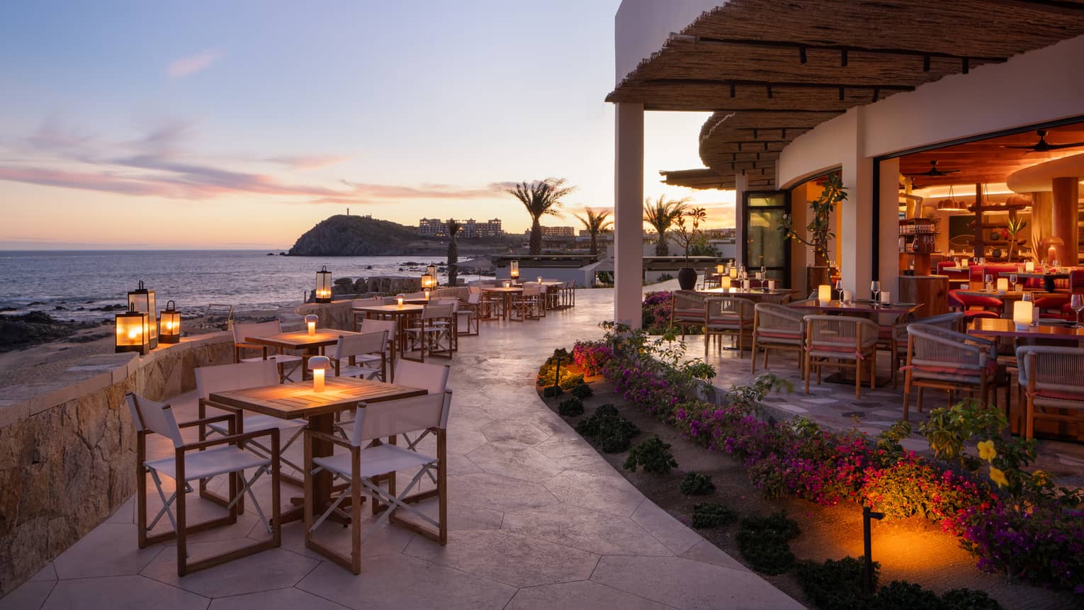 In the warm afterglow of sunset, lanterns light up a large, curved stone restaurant patio at the edge of the ocean.