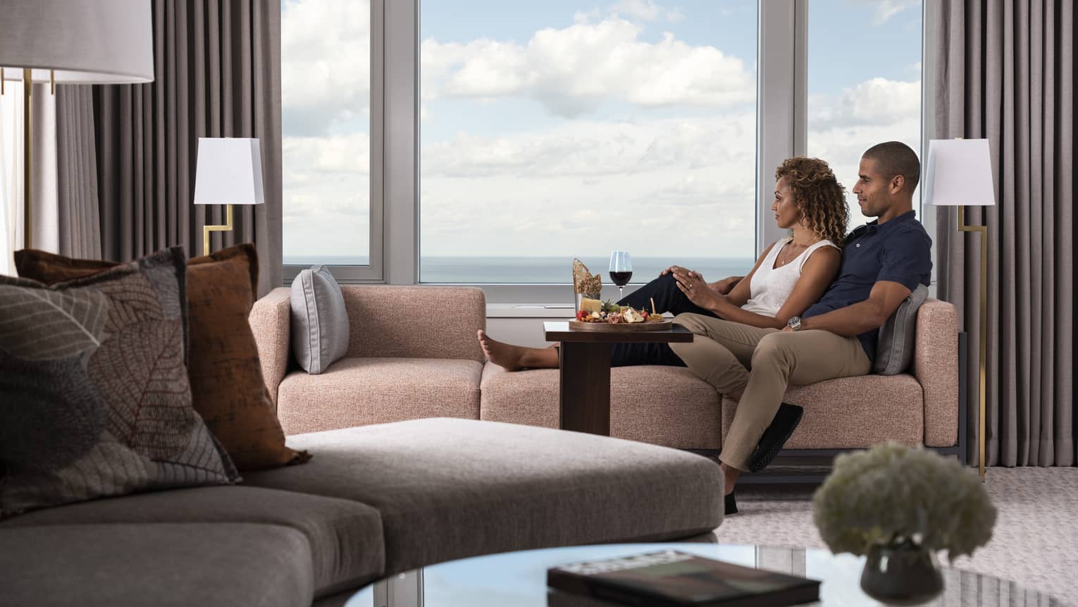 Couple lounging by windows on muted red tweed sofa flanked by standing floor lamps, lake view