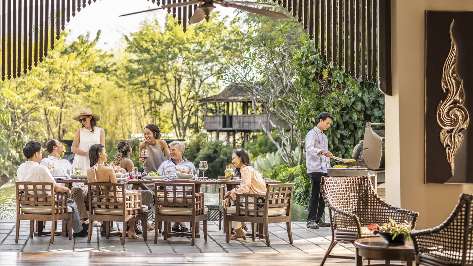 A smiling group around a rustic patio set with wine and appetizers, as a chef barbecues corn and light filters through trees.
