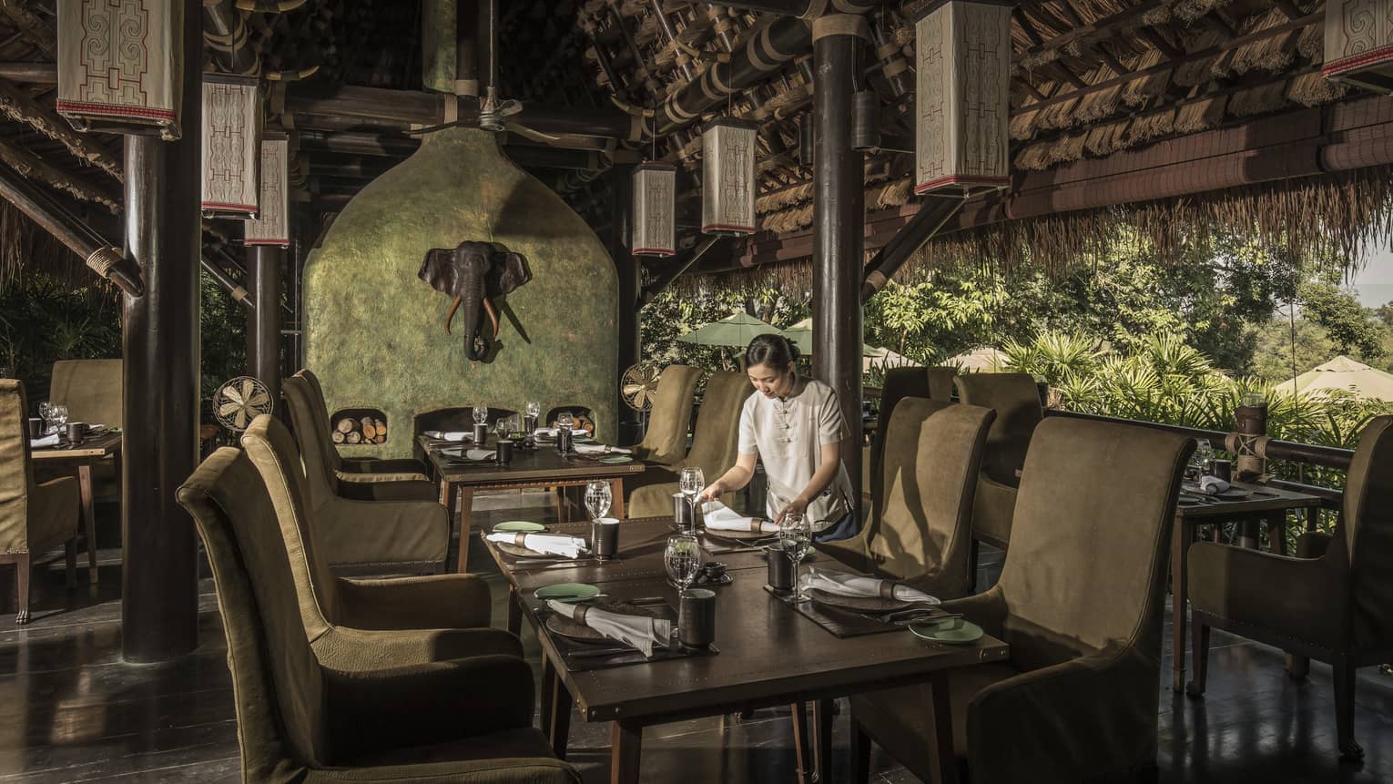 A woman sets the table in an open-air restaurant, natural-coloured furnishings, elephant head decoration on wall