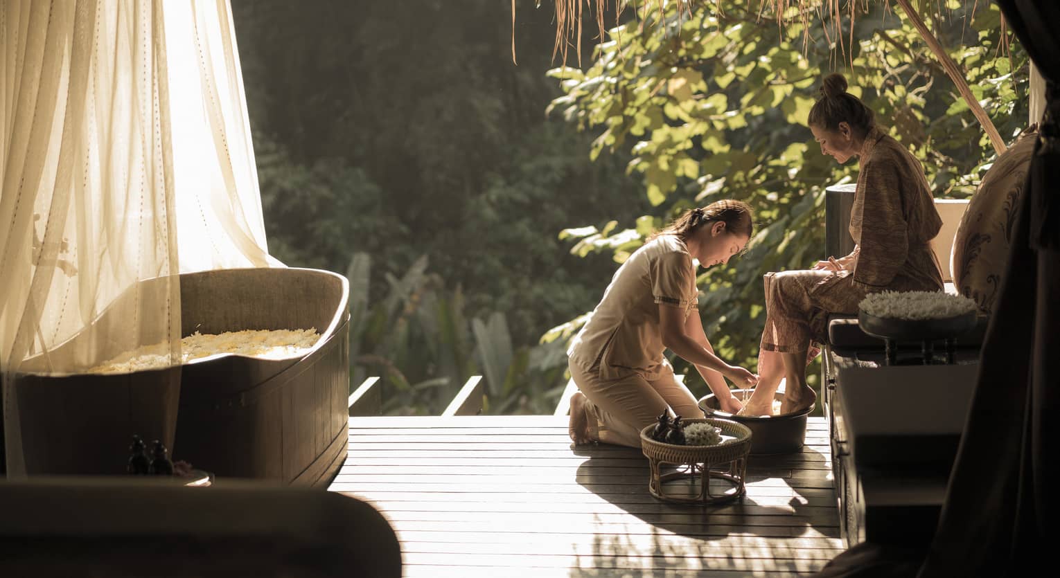 Spa technician kneeling, rinsing feet of woman seated, wearing spa robe under thatched roof