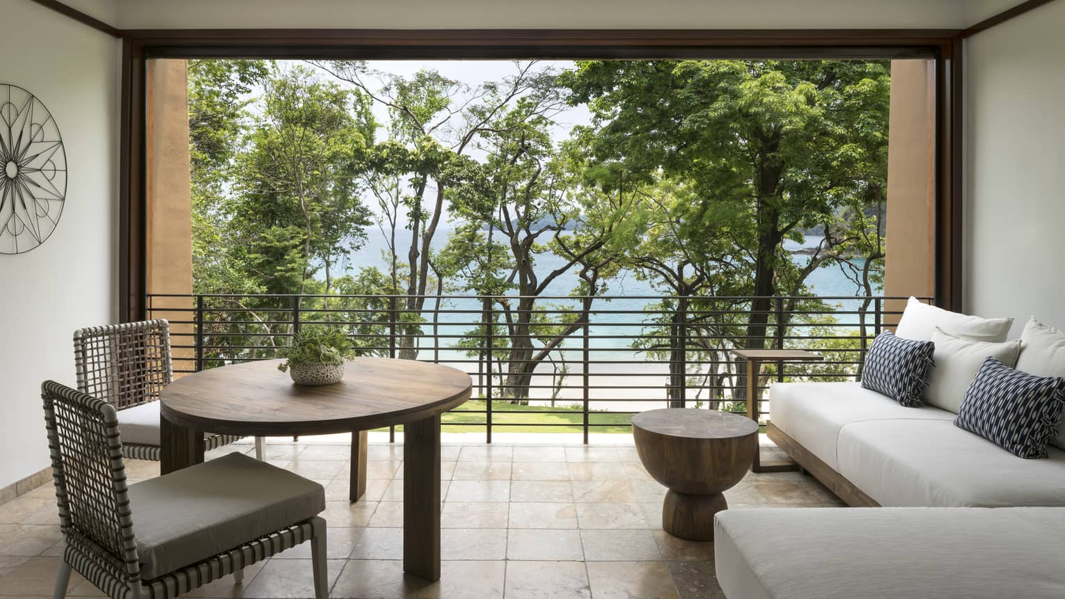 Terraza Room round dining table with two chairs, L-shaped sofa on tile floors, open wall to iron balcony, trees