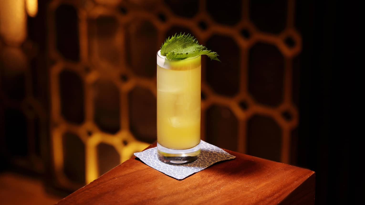 A yellow cocktail garnished with a green leaf sits on the corner of a wooden table