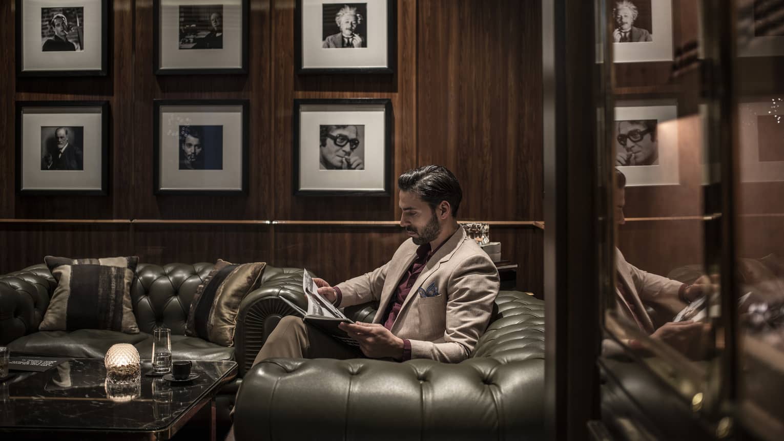 Person wearing tan suit and burgundy shirt sits on a black tufted leather sofa reading a magazine in a wood-panelled room