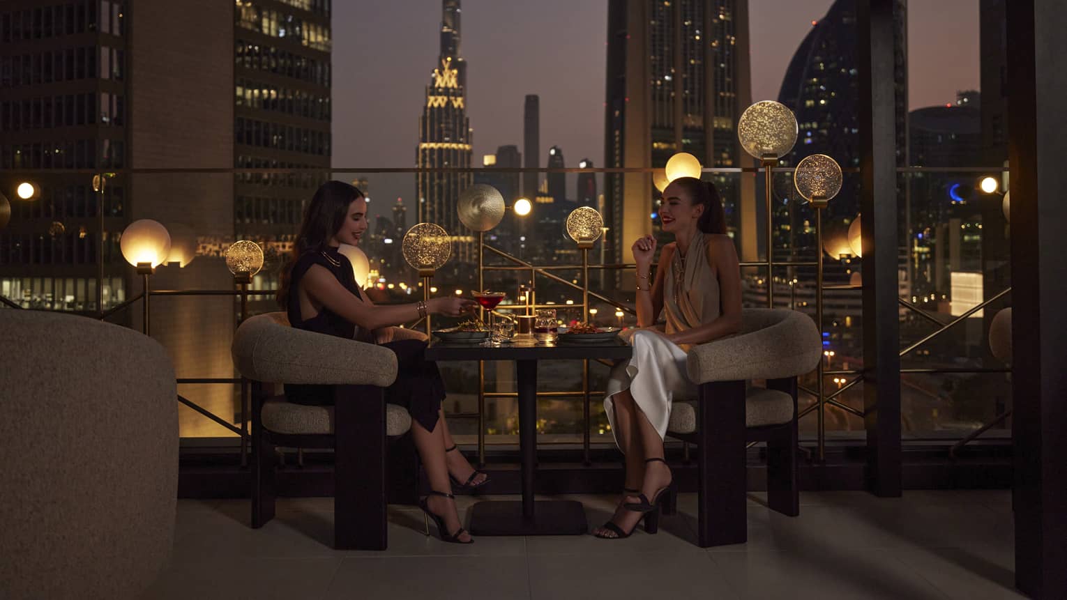 Two women sit at a cocktail table on an outdoor rooftop patio overlooking the Dubai skyline at night