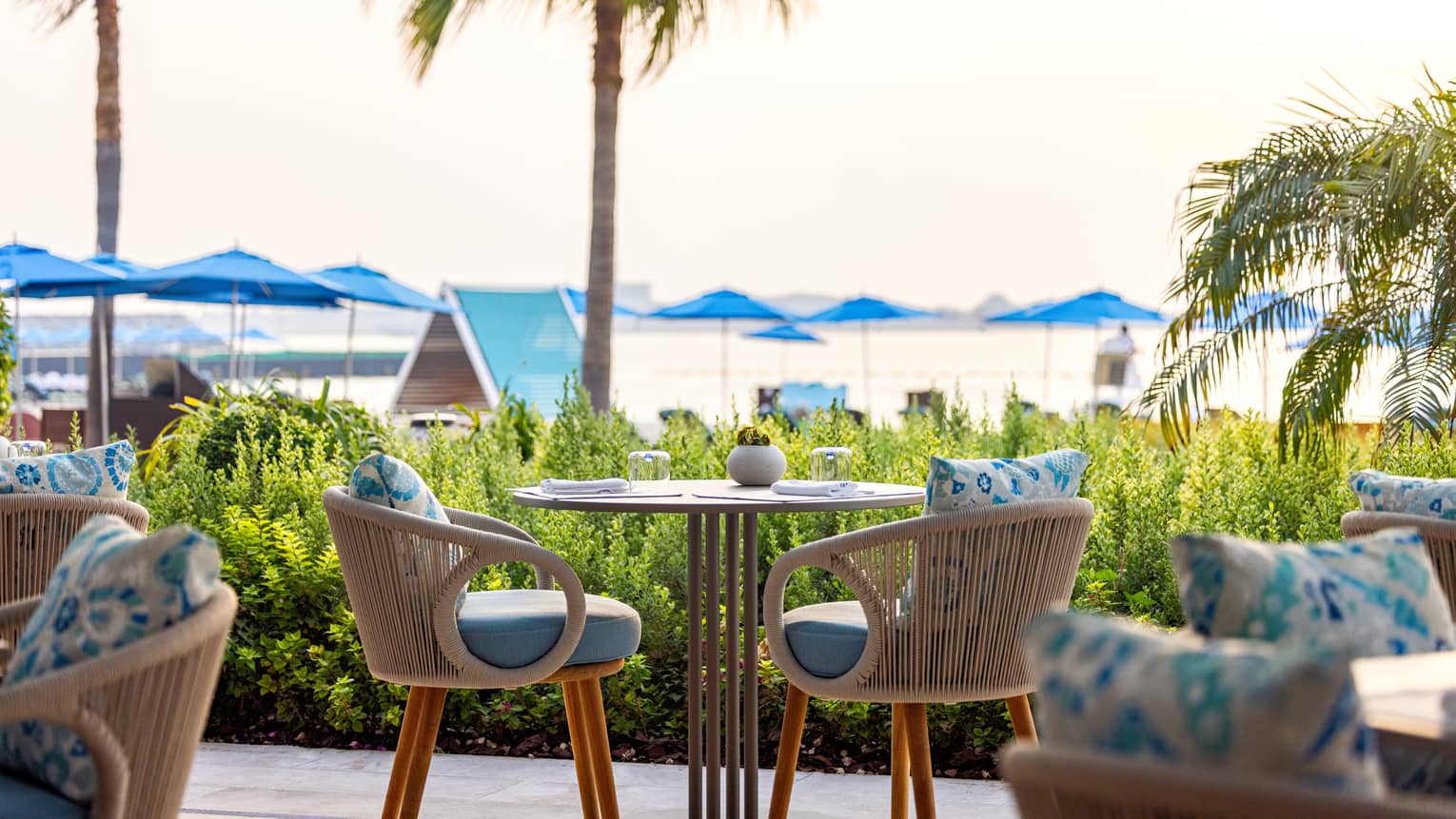 Small table for two set on an open-air terrace overlooking a lawn with palm trees and beach umbrellas in the distance