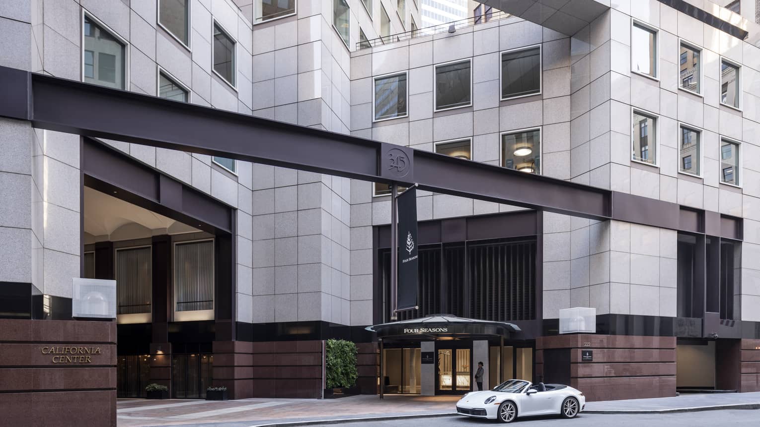 A white luxury sports caris parked in front of the massive Four Seasons hotel entrance, which is clad in pale grey granite.