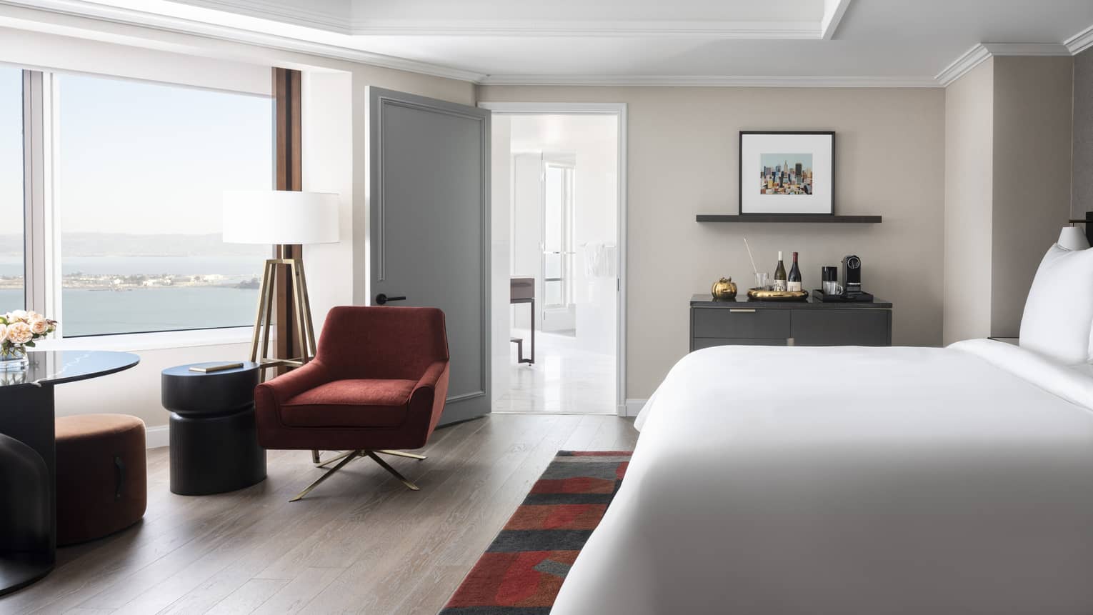 Modern hotel bedroom with light beige walls, white king bed, dark red arm chair, table, large window