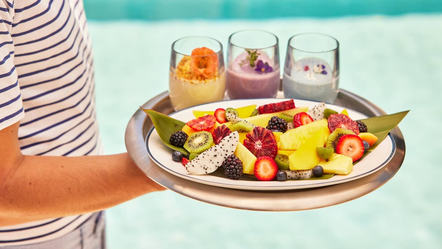 Person carrying tray of smoothies and fruit by pool