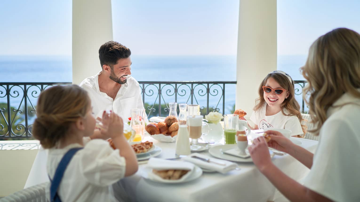 Family of four smiling while sitting at breakfast table overlooking the sea