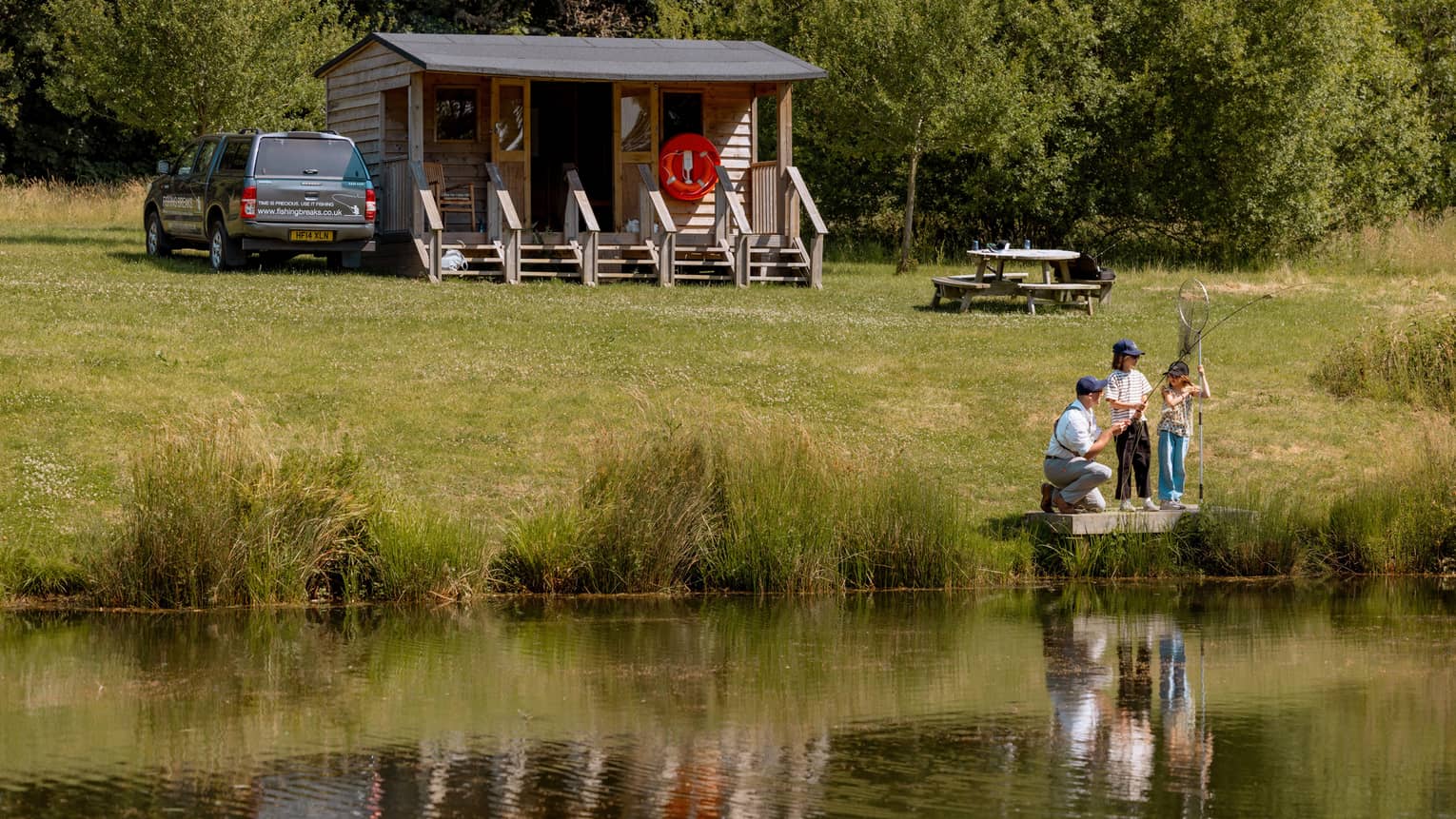 Three people fishing on small platform by lake with cabin-like building and picnic table in backdrop