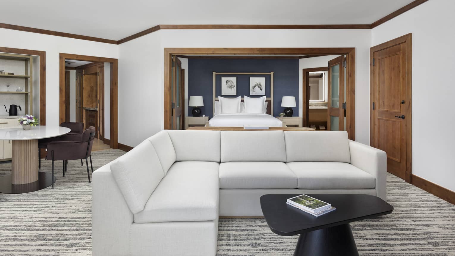 Open luxury suite with a light grey sectional sofa, black coffee table, table with two chairs and a view into the bedroom with a king bed 