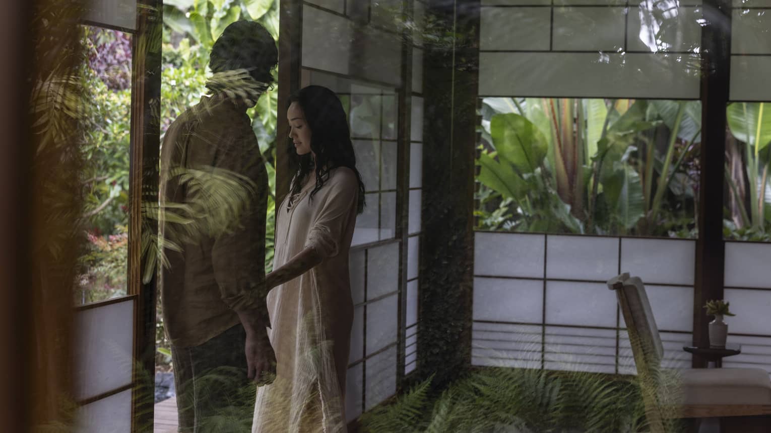 In a glass hut surrounded by abundant greenery, a couple stands at the open entrance. One places a hand on the other’s arm. 