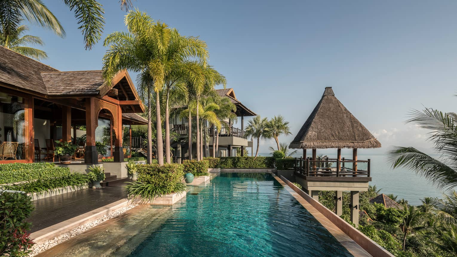 Long outdoor swimming pool under tall palm trees, thatched roof gazebo