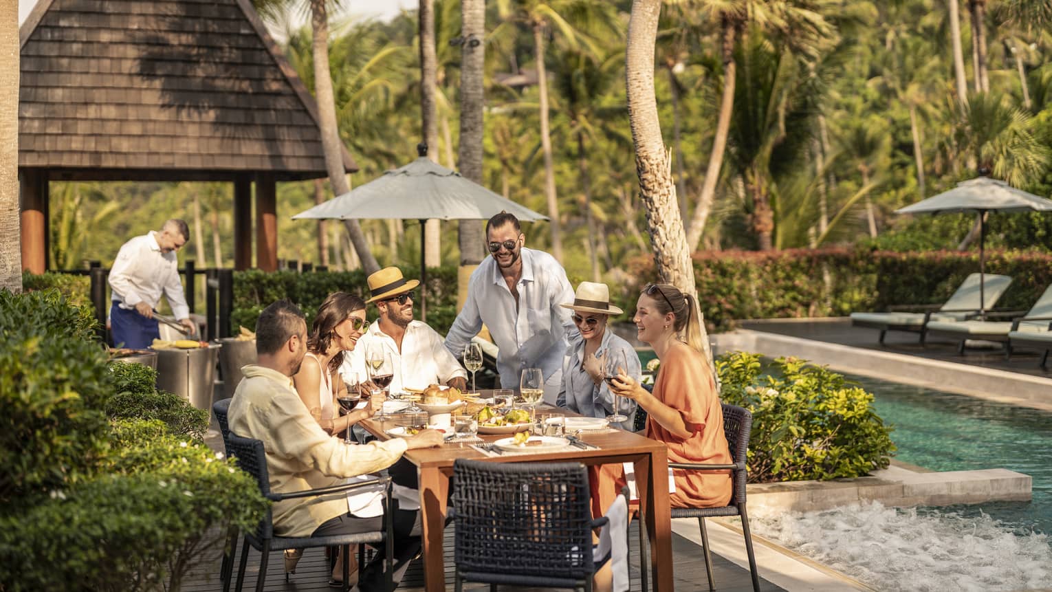 Group of laughing friends enjoy a poolside barbecue lunch
