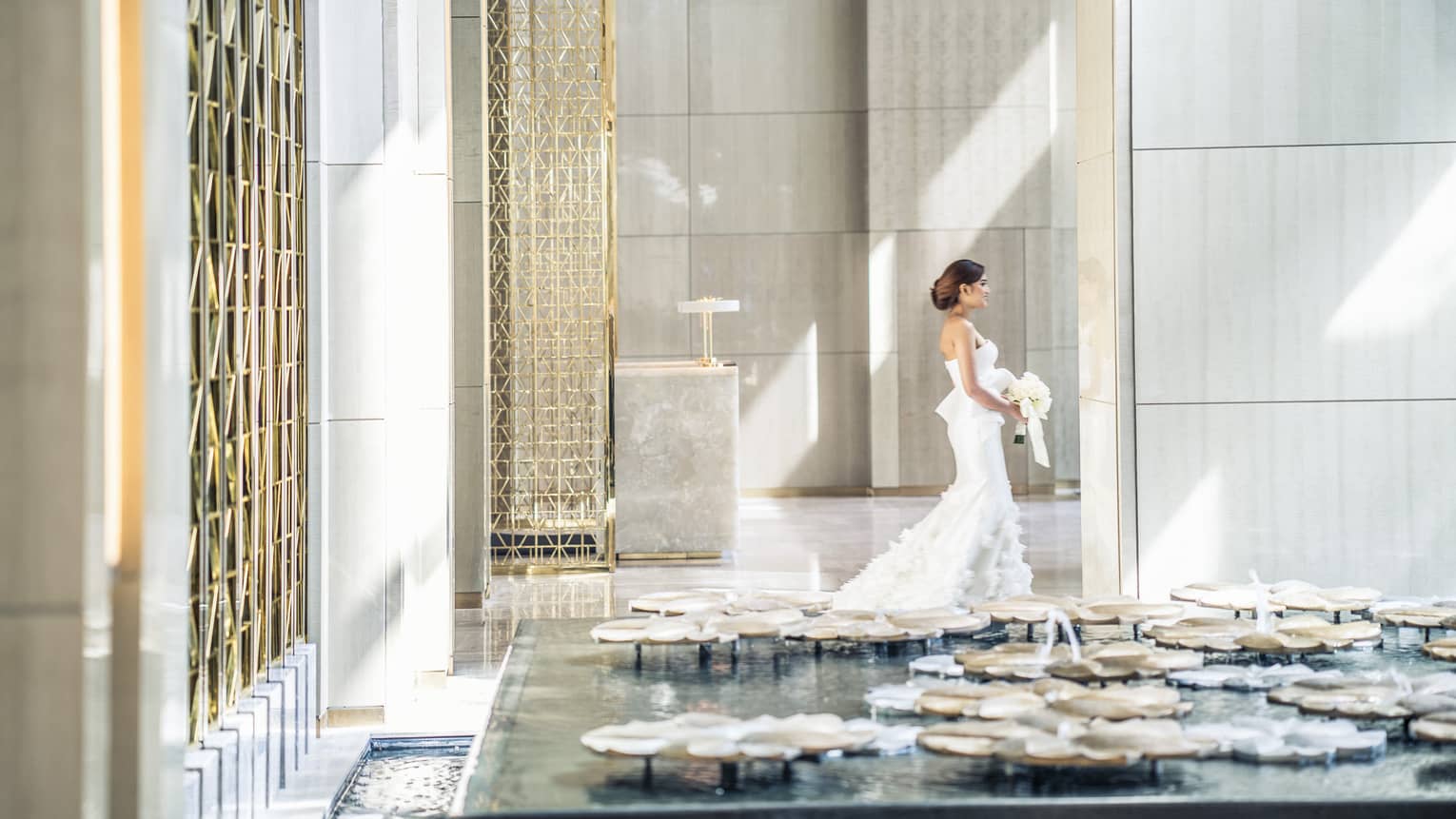 Sun rays illuminates a bride as she walks through a white walled lobby carrying her bouquet of white flowers
