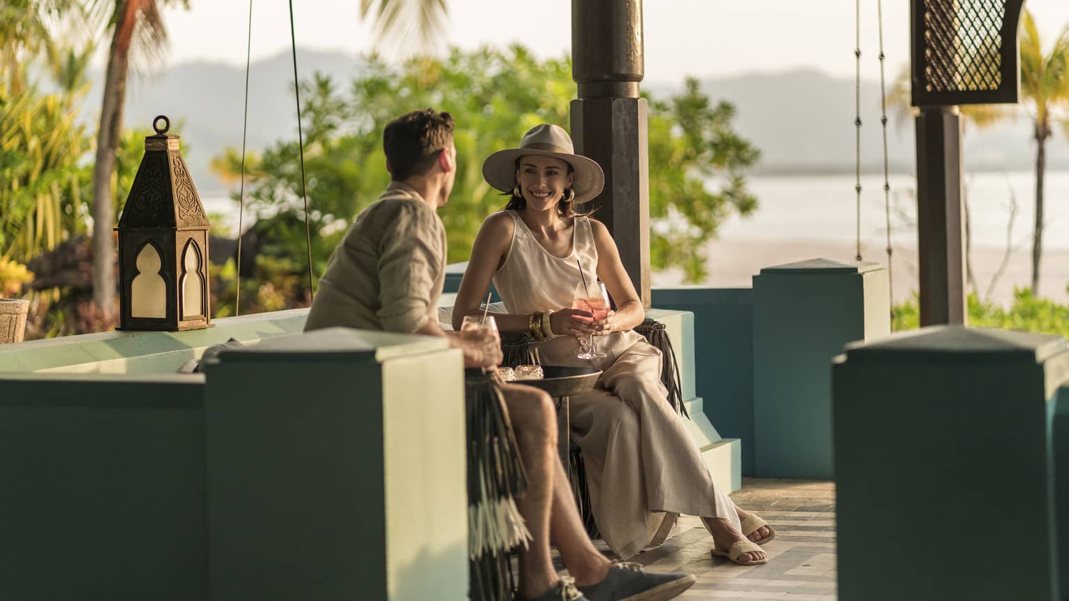 A man and woman are sitting in an outdoor Rhu Bar area overlooking the ocean holding cocktails