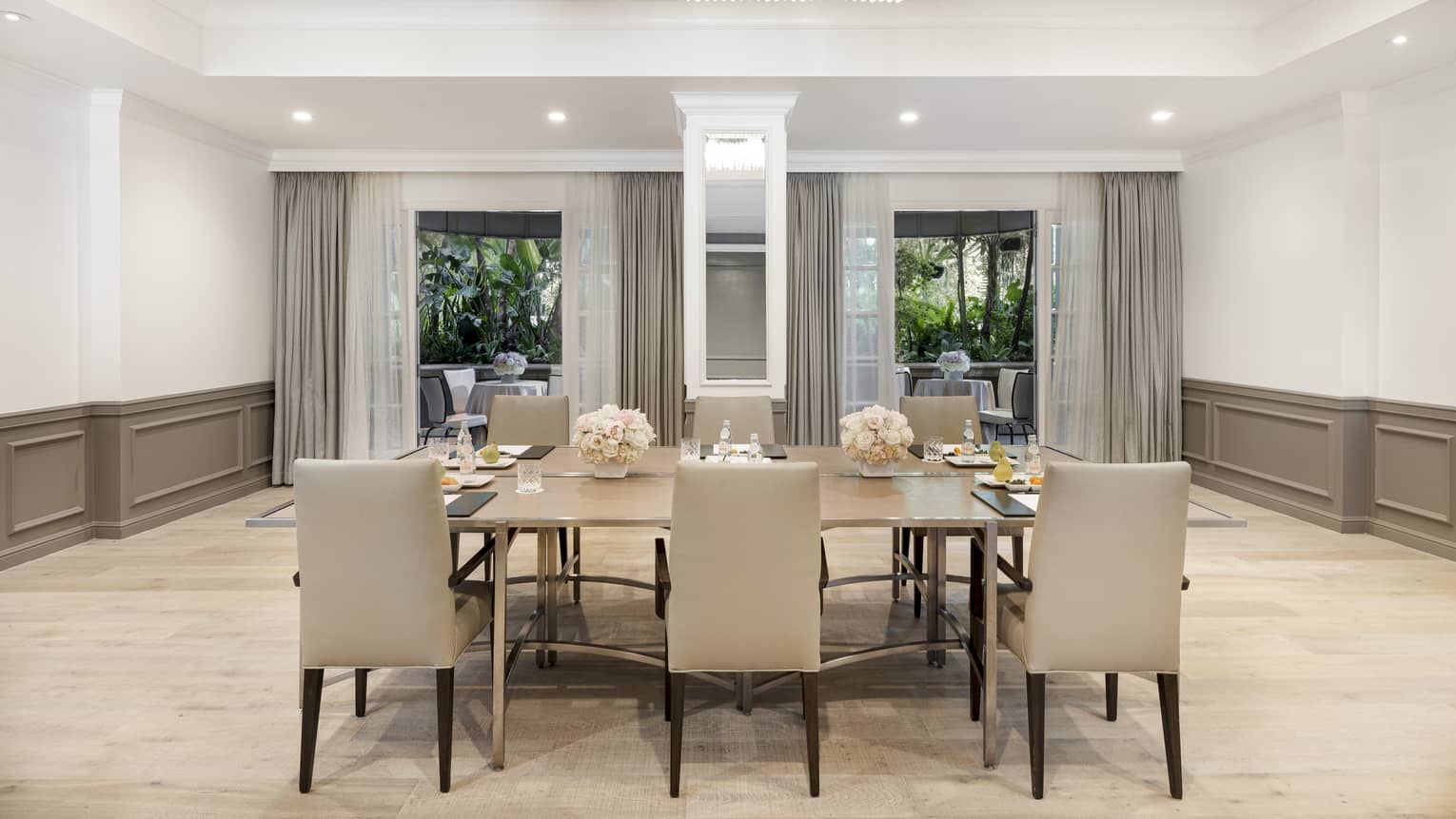 A large table with six chairs in a large room with beige accents and large windows.