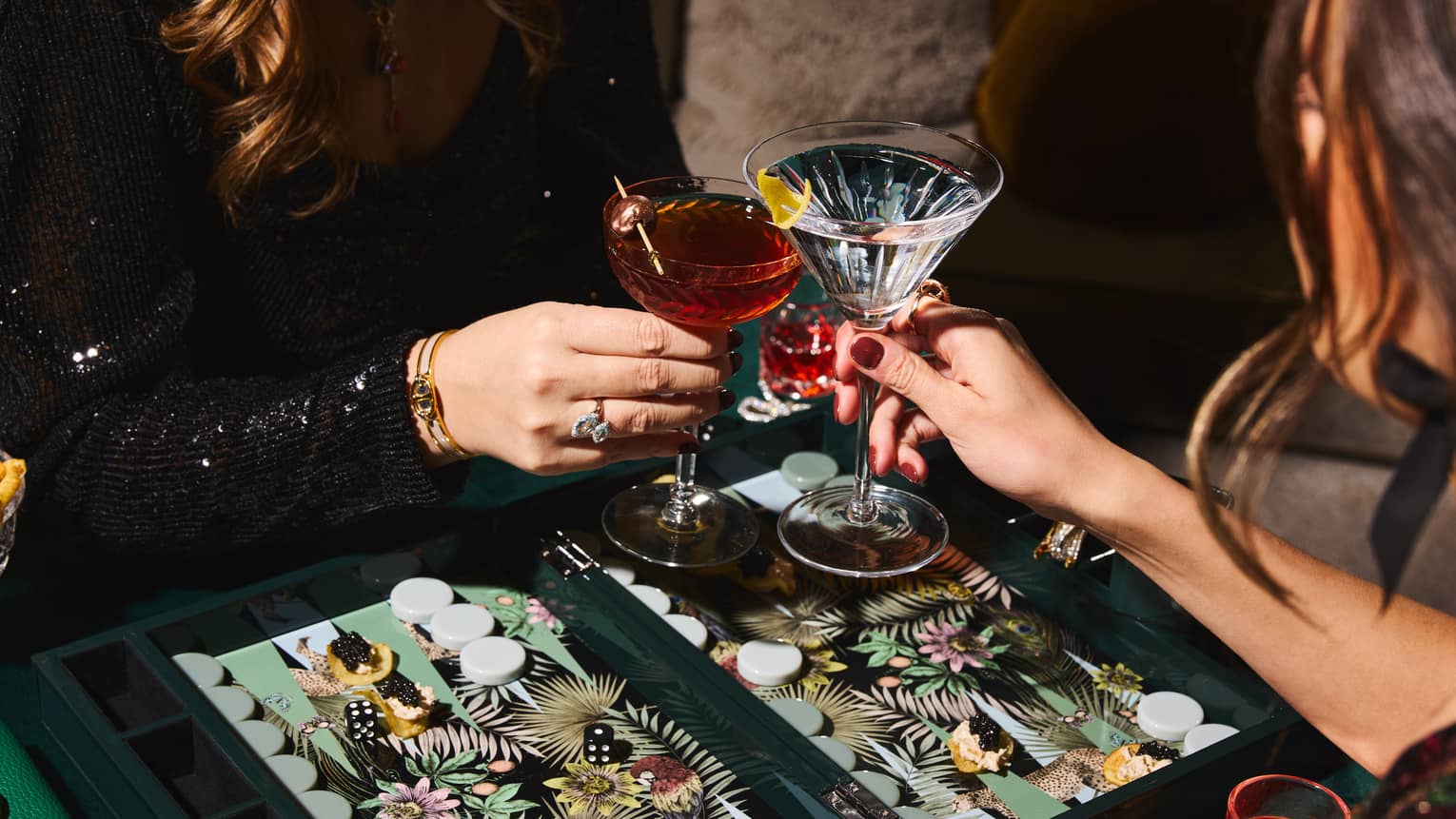 Two people toast their Martini glasses together over a colourful backgammon board