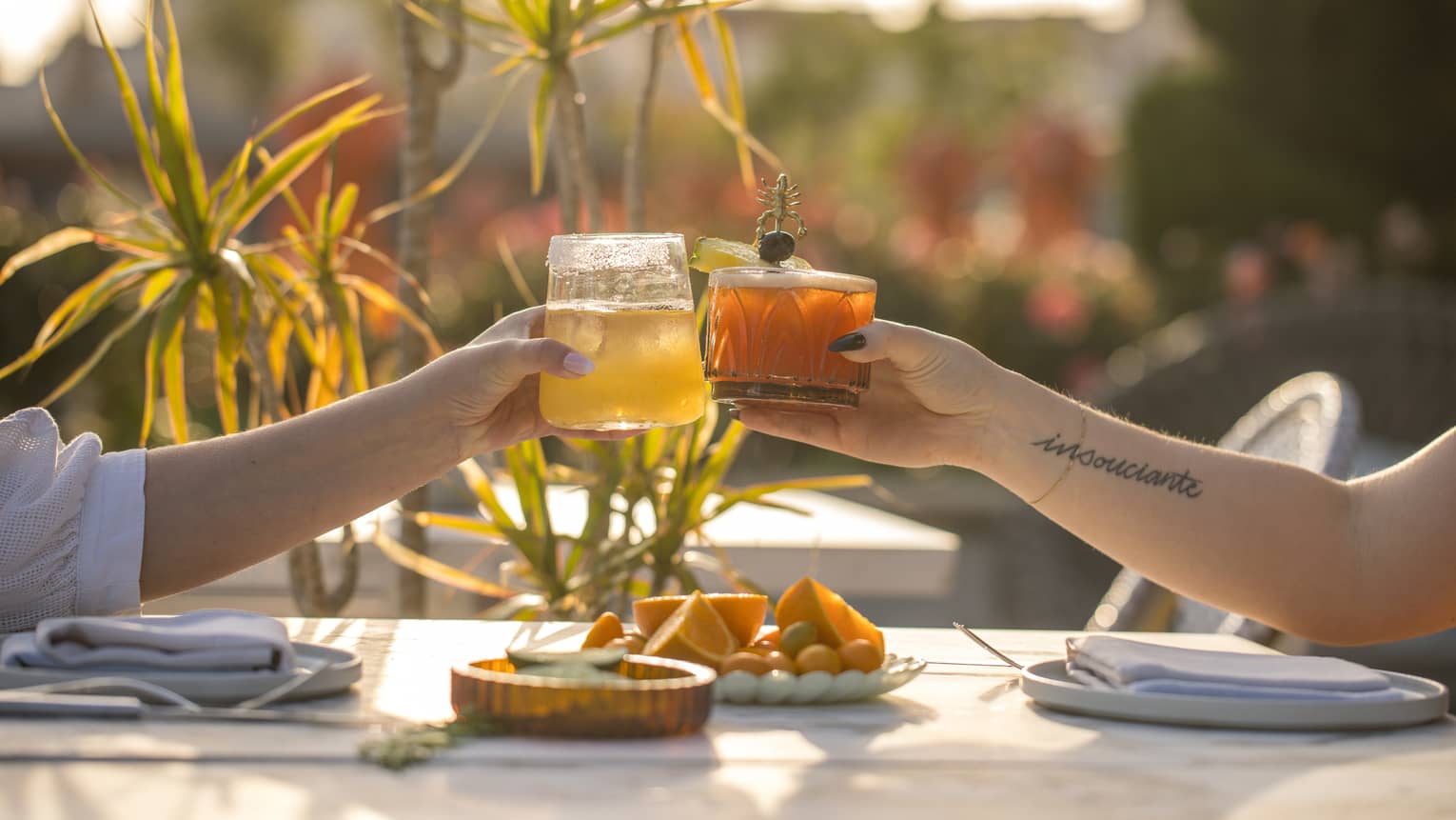 Two hands, each holding a cocktail, bring their glasses together over a sun-lit outdoor table