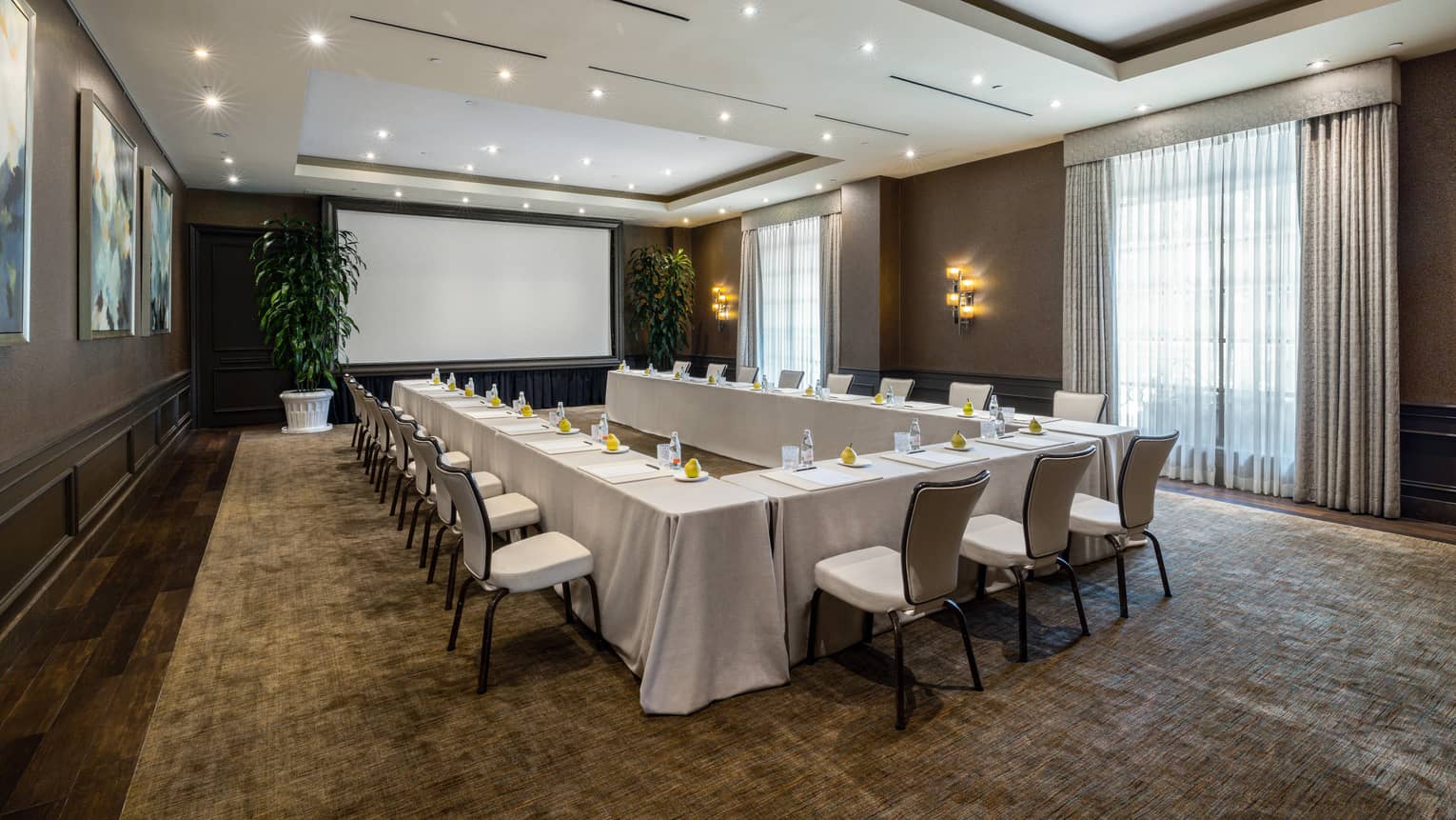 Meeting room set up U-shape style with cream-coloured linens, ambient lighting, projection screen framed by large potted plants, and floor-to-ceiling windows