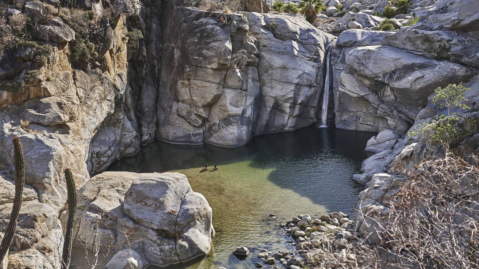 Two people wade in a small ocean lagoon surrounded by large cliffs, creating a serene and picturesque scene with clear water and rugged natural beauty.