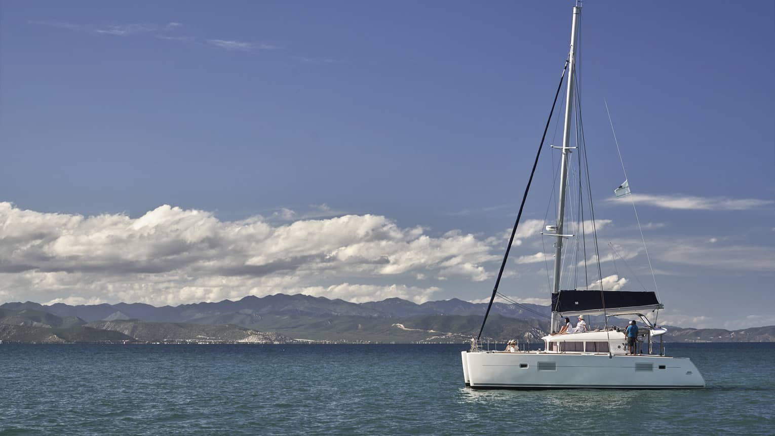 A catamaran on calm, blue water under a partly-cloudy sky with low green hills in the background and mountain ranges beyond.