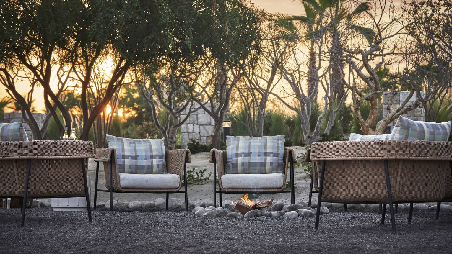 Four wood and cushioned chairs surrounding a small fire on the ground, the sun is setting behind a group of trees.