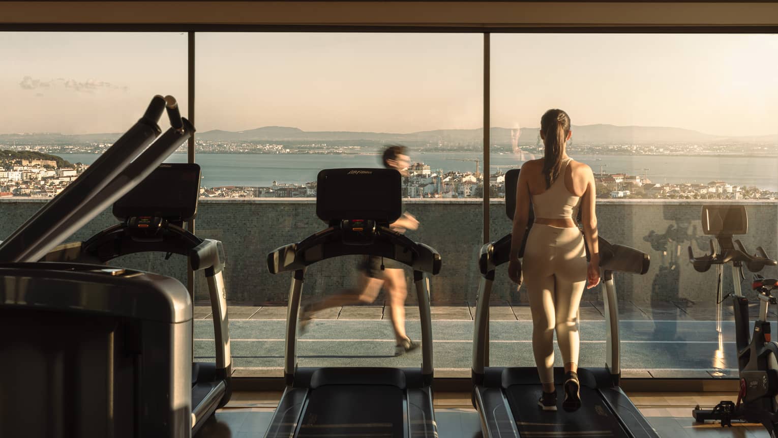 A person uses a treadmill facing floor-to-ceiling windows in a rooftop fitness centre. Another person is running by on the track outside, and the coastline and water are visible in the background.
