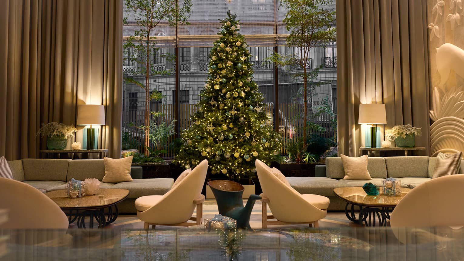 A Christmas tree decorated with gold and teal ornaments stands before a large window in a neutral-coloured seating area.