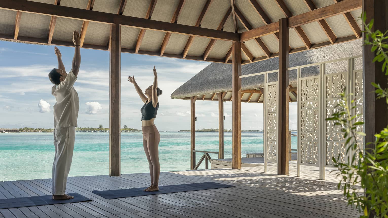 Two people practising yoga on a shaded deck overlooking turquoise ocean water at a tropical island retreat