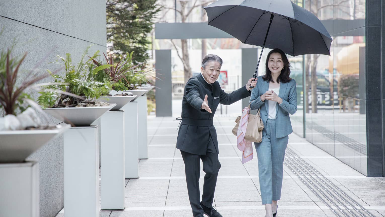 Staff member leads smiling guest down walkway while sheltering them with large black umbrella