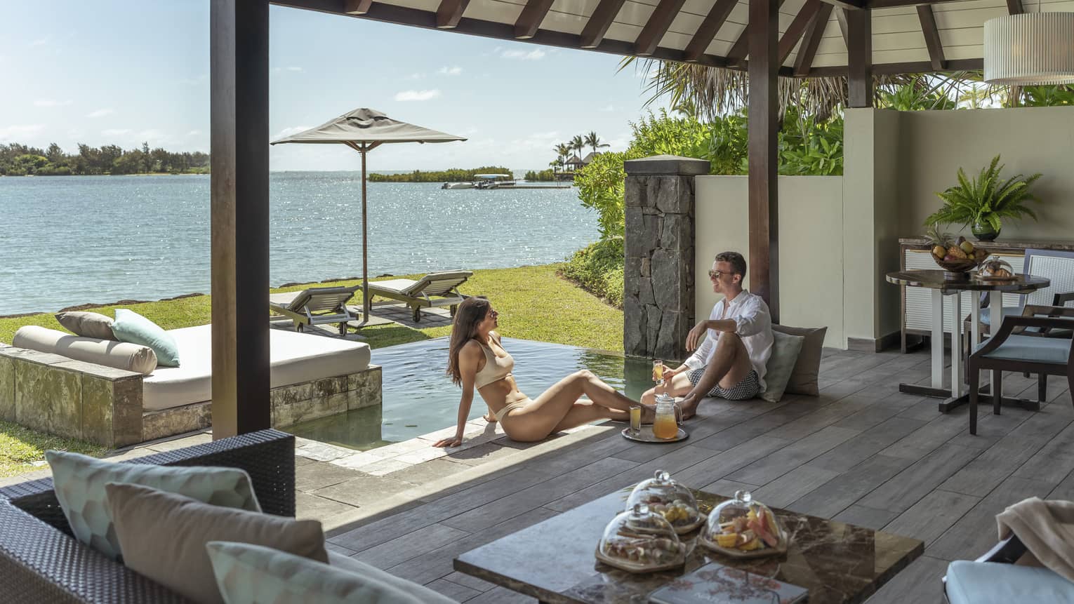 Man and woman sit on wooden terrace next to small pool, sea view
