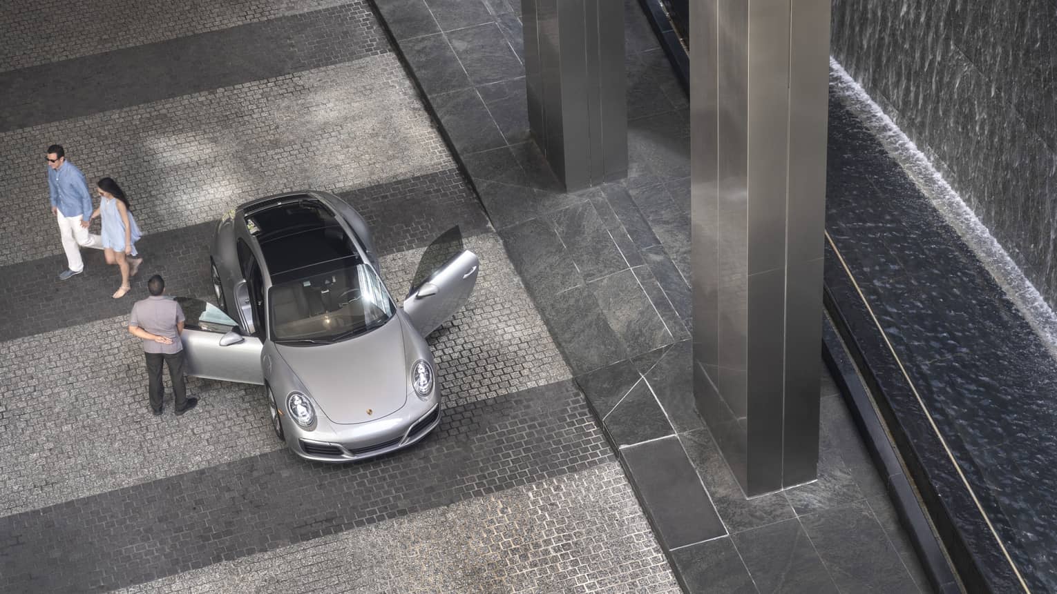Aerial view of hotel staff opening door of luxury silver car on driveway, couple walking in background