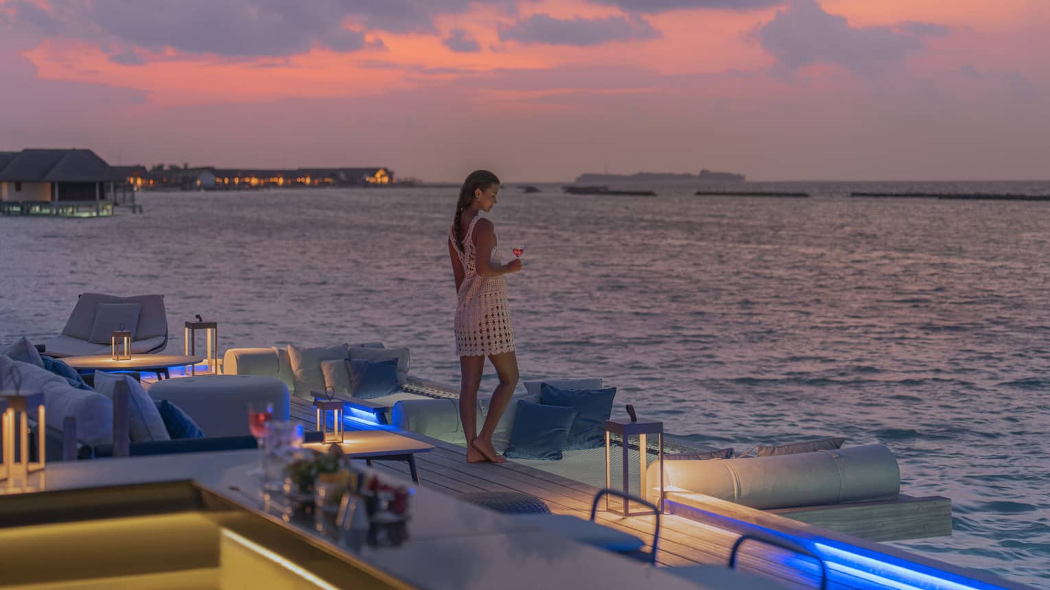 Woman holding cocktail stands at edge of Seabar overlooking the water at sunset