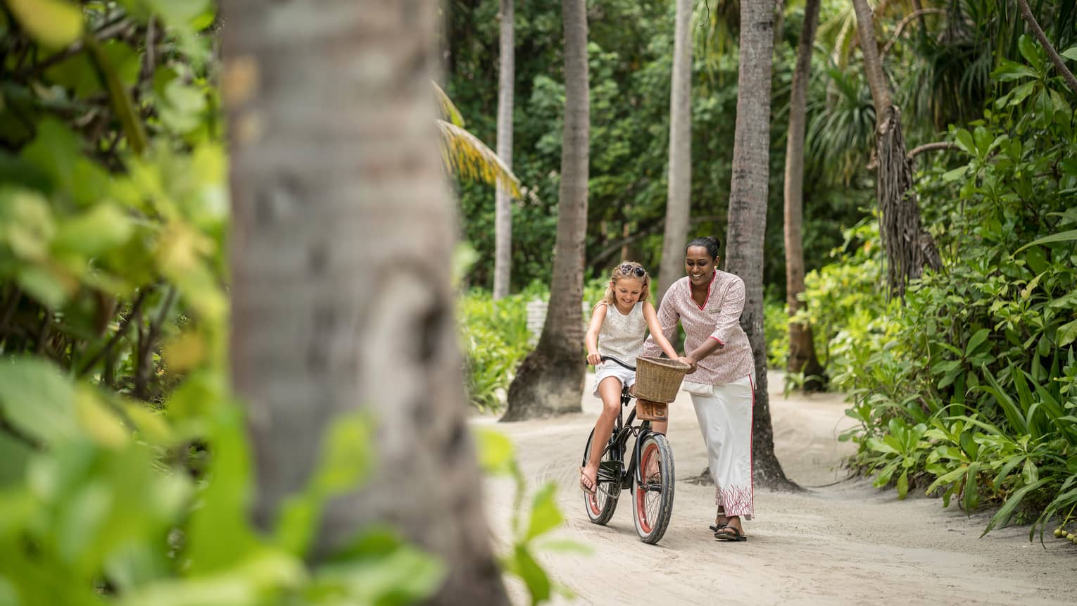 Hotel staff helps young girl ride bicycle with basket down resort path past tropical forest