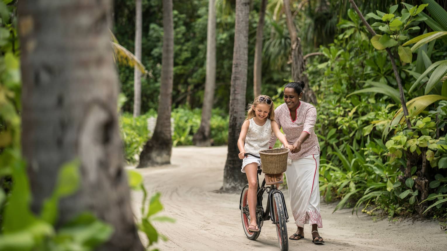 Hotel staff helps young girl ride bicycle with woven basket on tropical resort path