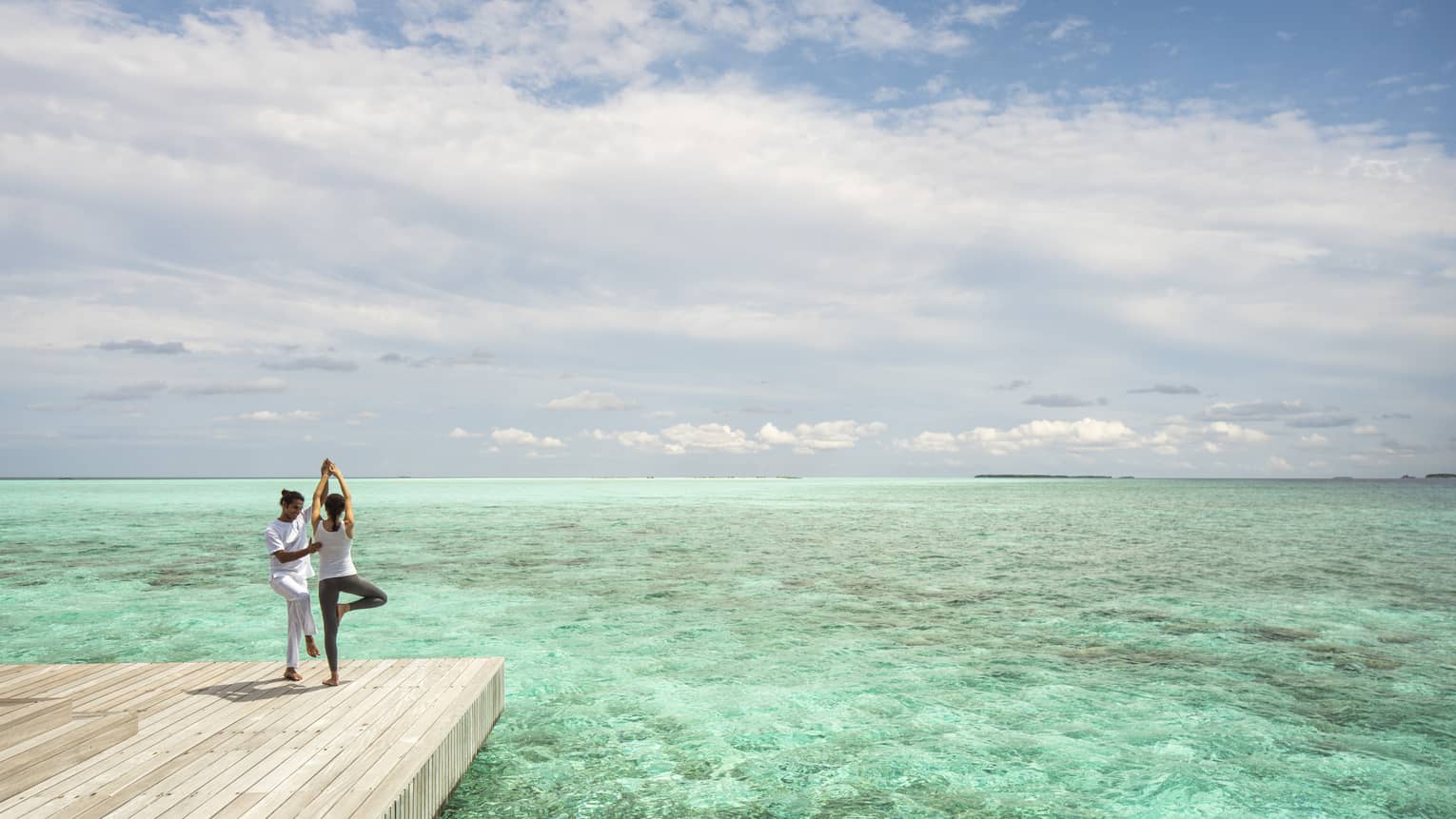 Yoga instructor helps woman with tree position on wooden pier beside ocean