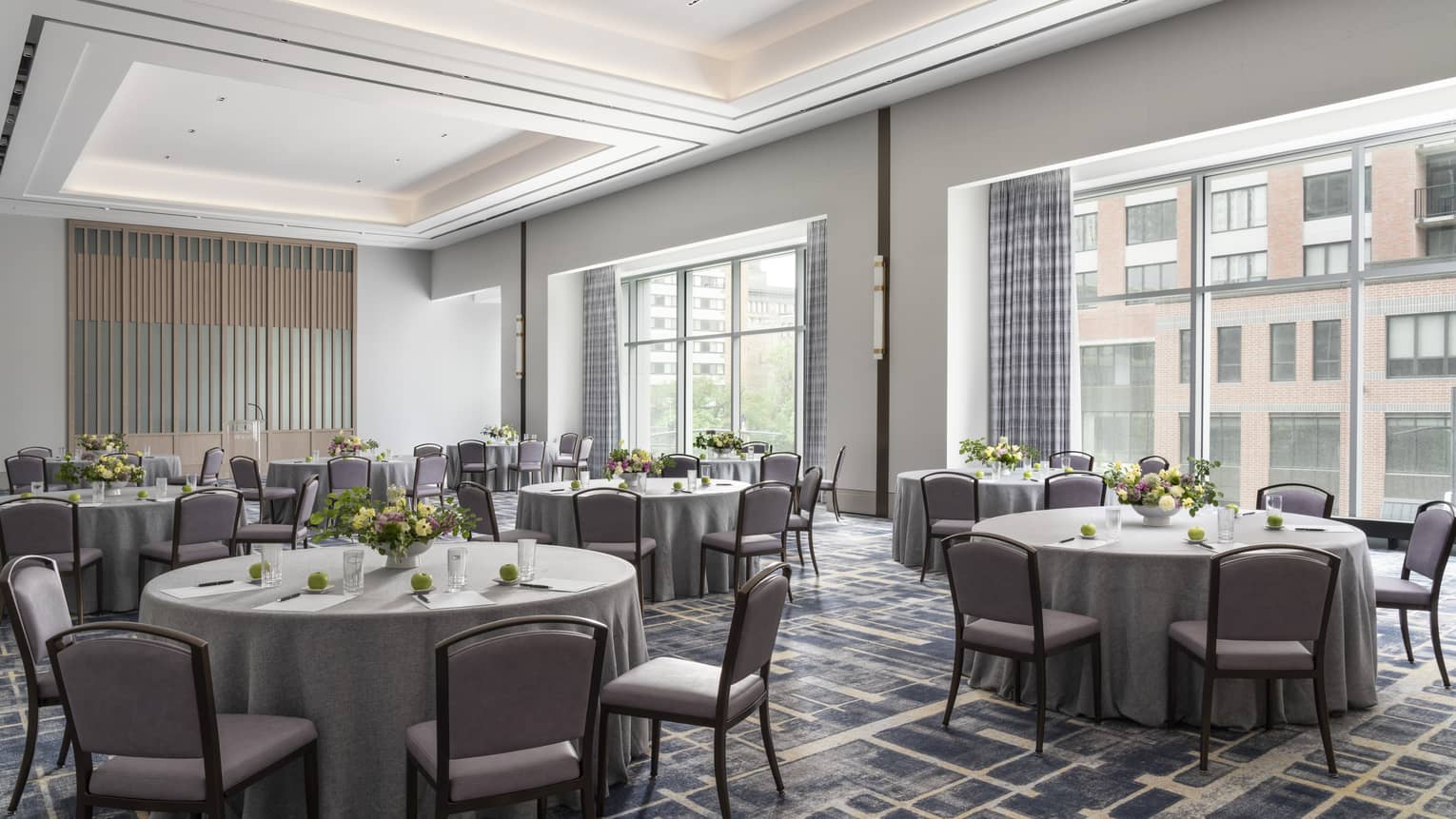 A ballroom with round tables covered in grey table cloth and yellow flower, with large windows in the room.
