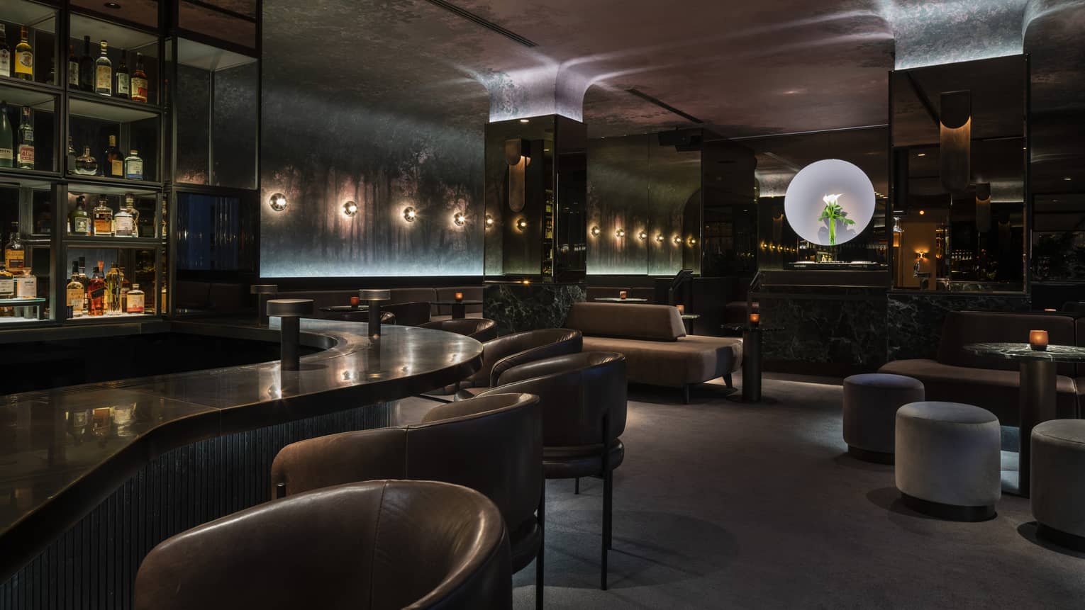 Chic lounge with curved bar, leather chairs and stools and moody lighting, walls and ceiling covered in a dark forest mural. 