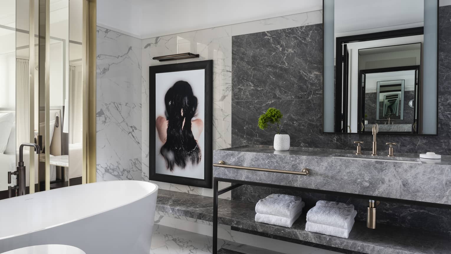 A freestanding tub, grey marble vanity and a framed work of art are set beside glass doors that open to a bedroom